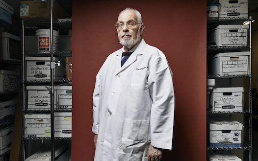 Portrait of David Egilman, wearing a lab coat and standing in front of fileboxes